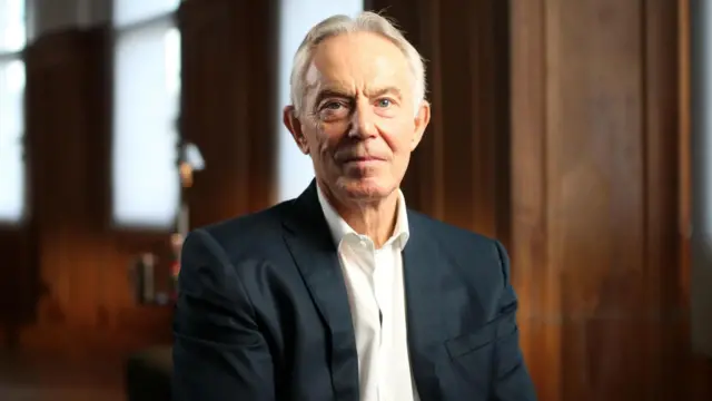 Tony Blair olha para a câmera. Ele está sentado, vestindo um terno azul-marinho e uma camisa branca com a gola aberta.