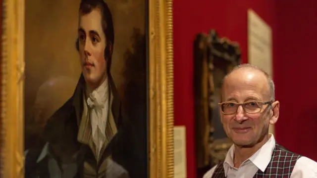 El Dr. Bill Zachs, calvo y con chaleco a cuadros rojos, amarillos y oscuros, camisa blanca y gafas color mostaza, se encuentra junto al retrato redescubierto de Robert Burns realizado por Henry Raeburn, que se exhibe en las Galerías Nacionales de Escocia.