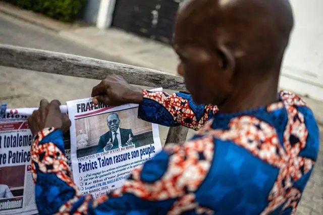 Un homme vêtu d'un imprimé traditionnel ouest-africain bleu et orange tient un journal dont la une montre une photo de Patrice Talon, le président béninois.