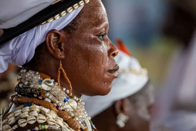 Une femme est prise en photo de l'épaule à la tête. Elle porte de nombreuses perles autour du cou et aux épaules, son visage est rempli de sueur et elle semble plongée dans la méditation.