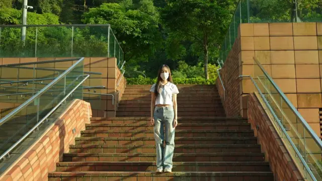 一名戴口罩的女性站在紅磚階梯中央，背景是綠樹環繞的戶外環境。