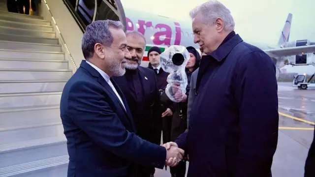 Двое мужчин пожимают друг другу руки у трапа самолета, на борту которого изображен флаг Ирана.