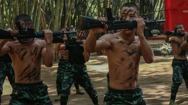 ဝါးရုံတွေဝန်းရံထားတဲ့ နေရာမှာ စစ်သင်တန်းဆင်း တပ်သားအမျိုးသားရော အမျိုးသမီးတွေပါ သေနတ်ကိုကိုင်ကာ စစ်ရေးလေ့ကျင့်သရုပ်ပြနေကြပြီး အခမ်းအနားအဖြစ် အနီရောင်နောက်ခံပိတ်စသုံးကာ ပြင်ဆင်ထားပါတယ် 