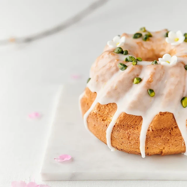 Na białej marmurowej podstawce stoi okrągła babka pieczona w formie z kominem. Ciasto ma złocistobrązową powierzchnię i jest obficie polane białym lukrem, który spływa po bokach w długich smużkach. Wierzch babki ozdobiony jest zielonymi pistacjami oraz małymi białymi dekoracjami przypominającymi kwiaty. Tło jest jasne i rozmyte, a wokół na powierzchni leżą pojedyncze różowe płatki.