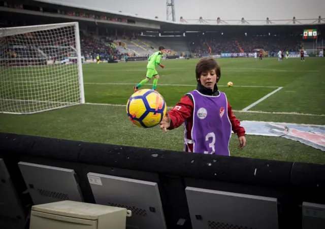 Arda Güler top toplayıcı olarak görev yaptığı maçta, elindeki topu saha kenarına atıyor. Arkasında maç devam ediyor.