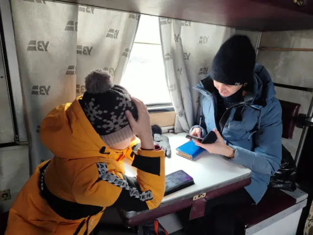 Дитина у жовтій куртці сидить у плацкартному вагоні навпроти жінки у синій куртці 