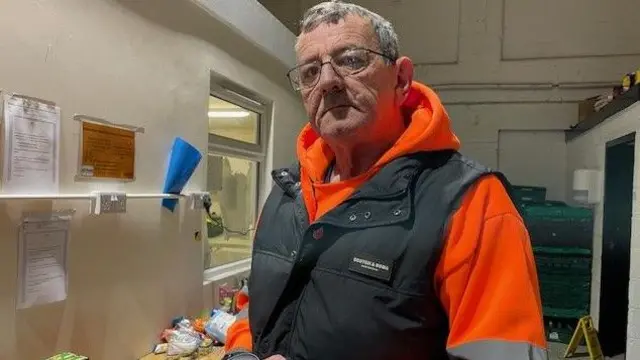 Un hombre con un chaleco negro sobre una sudadera naranja con capucha sostiene una lata en la mano. Está de pie en una pequeña habitación.