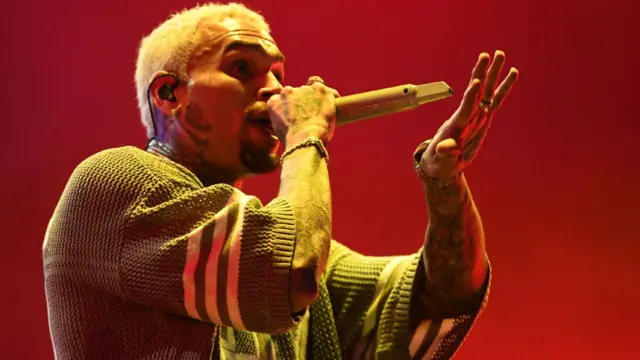 US musician Chris Brown, dressed in a khaki-knit top with tattoos on his arms and neck and short blonde hair, holds a mic as he sings. The background is red.