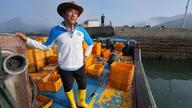 Um homem usando chapéu, jaqueta clara e botas amarelas brilhantes está no convés de um barco atracado, ao lado de várias caixas amarelas.