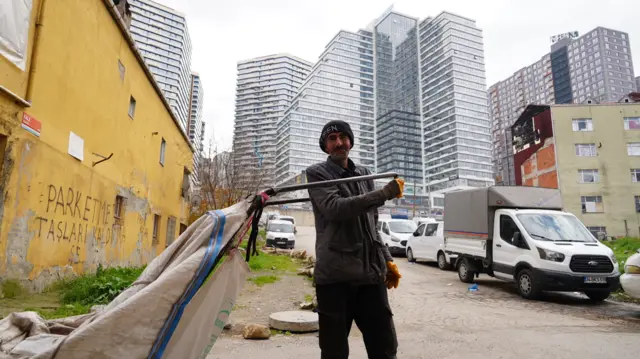 Semih Hazer çekçek ile poz veriyor. Bulunduğu sokakta eski binalar, arkasında ise yüksek katlı bir rezidans görünüyor.