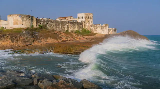 Vista lateral do exterior do Castelo de Cabo Corso com as ondas do Atlântico quebrando na praia abaixo das muralhas do edifício.