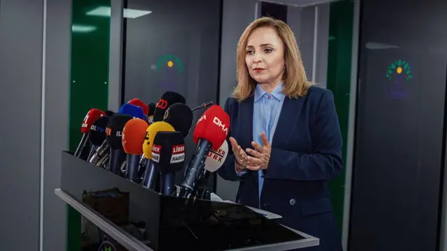 DEM Parti sözcüsü Ayşegül Doğan bir kürsüde konuşuyor, önünde çok sayıda mikrofon var. üzerinde açık mavi gömlek ve lacivert bir ceket var.