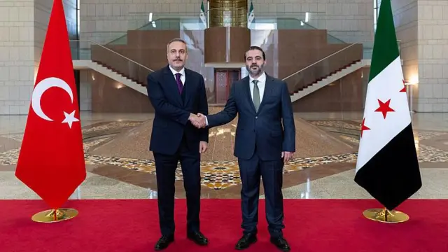 Hakan Fidan Şam'da Suriye Dışişleri Bakanı Esad Hasan Şeybani ile bayraklar önünde tokalaşıyor