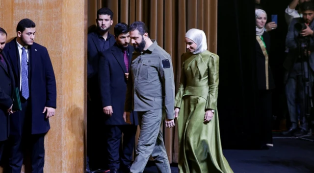 Ahmed Şara'y ve yeşil kıyafeti ile lila rengi başörtüsüyle eşi Latife el-Durubi sahnede yürüyor. Yakınlarında korumalar var.