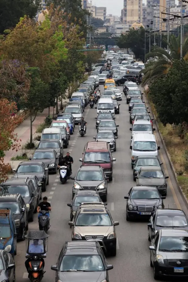多層階のビル群を背に、3車線の道路を埋め尽くす車列が手前に向かっている。合間を走るバイクもいる
