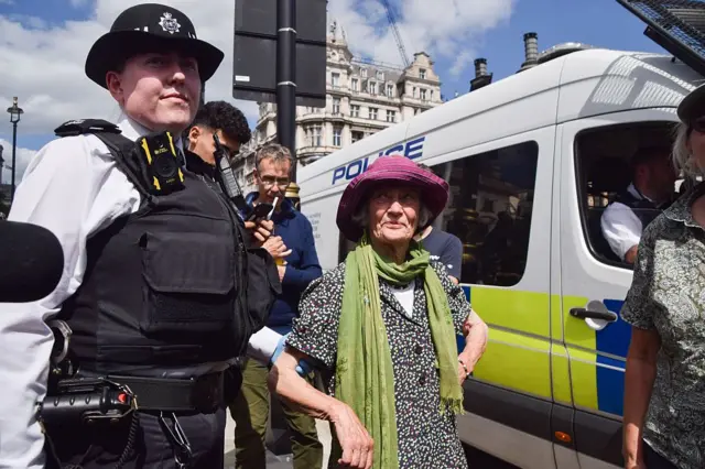 9 Ağustos'ta Londra'da düzenlenen eylemde gözaltına alınan bir kadın. Bir polis memuru kolundan tutuyor. Kadın yeşil bir kaşkol ve pembe bir şapka takıyor.