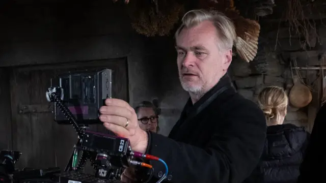 O diretor Christopher Nolan veste uma jaqueta preta e está no centro da imagem, olhando através de uma câmera IMAX