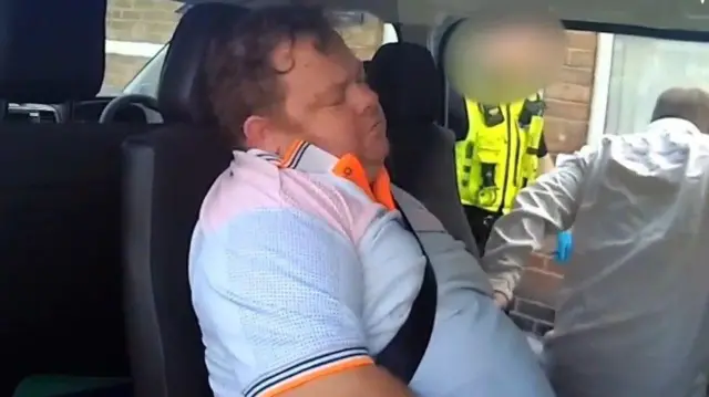 A man in a white and orange polo top pretending to have a disability in the back of a police car.