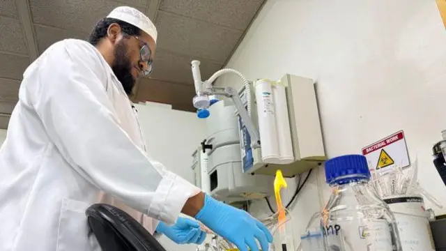 Prof Abdullah Ely ree ni yara iṣẹ iwadii ati ayẹwo , o wọ aṣọ funfun pẹlu ibọwọ alawọ aró.