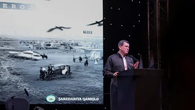 Abdi bir kürsüde konuşuyor arkasında duvara bir resim yansıtılmış, siyah beyaz bir şehri gösteriyor.