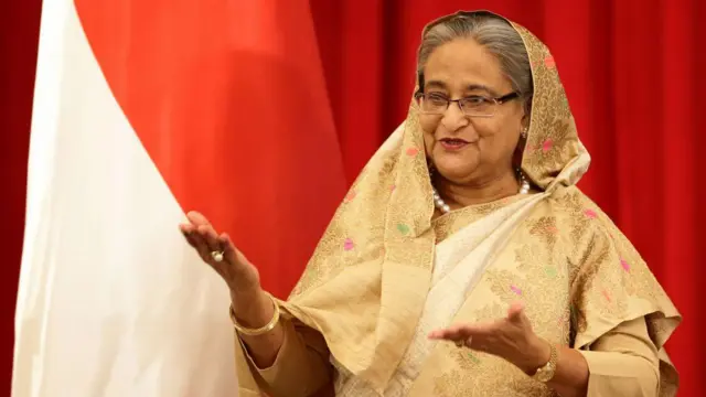 সোনালী রংয়ের শাড়ি পড়া শেখ হাসিনা দুই হাত সামনে বাড়িয়ে হাসিমুখে কথা বলছেন
