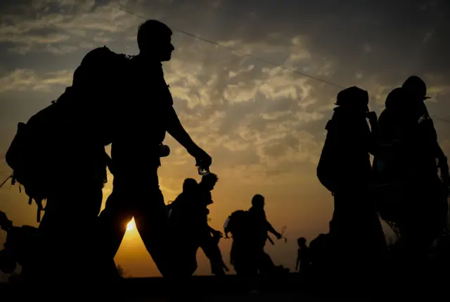 A group of people carrying lugguage on their backs,  walking before a sunset.