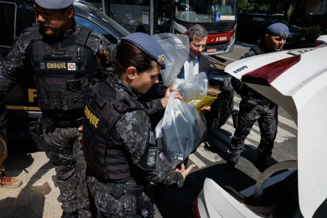 Policial coloca saco com material apreendido no porta-mala de carro; ela é rodeada por outros agentes