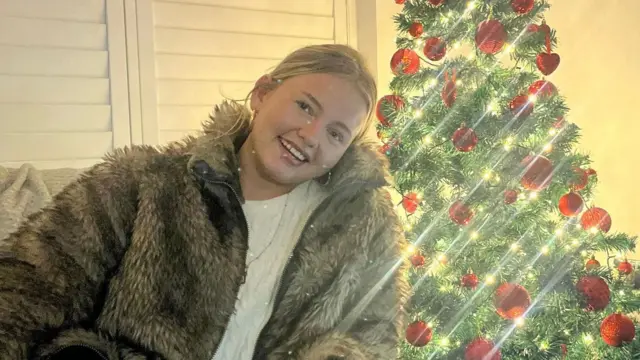 A woman with blonde hair in a fur jacket sitting in front of a Christmas tree with red baubles.