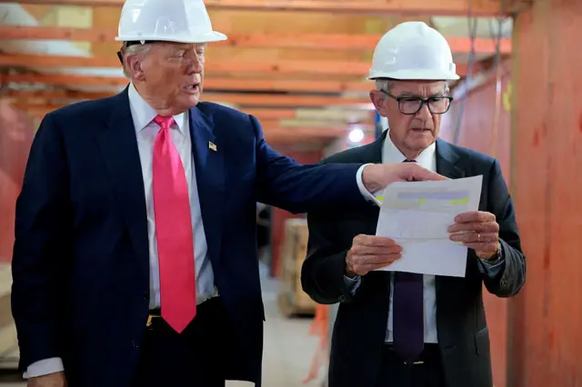 Donald Trump (i) señala algo en el documento que sujeta y mira Jerome Powell. Ambos llevan casco de obra y traje de chaqueta y corbata. 