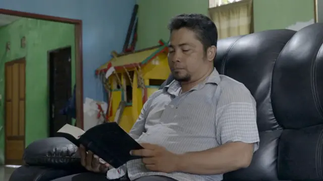 Sajie sedang duduk sambil membaca alkitab, di sudut rumahnya terdapat rumah Patahu. 