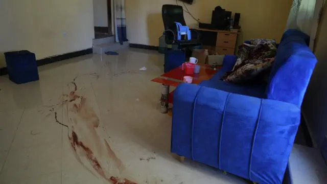 Aworan yii ṣafihan ẹjẹ to fẹ̀ lọ nilẹẹlẹ yara naa, aga alawọ eeru lo wa ni yara ọhun, tabili meji wa nibẹ pẹlu aga mi-in.