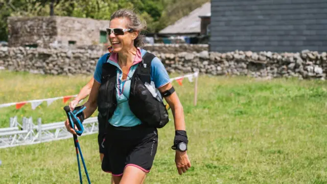 Anna Troup, que viste ropa de trail running que incluye pantalones cortos, mochila para correr, bastones, gafas de sol y un reloj deportivo, sonríe al llegar a un punto de descanso durante la Summer Spine Race.