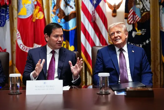 Rubio e Trump sentados diante de mesa, em frente a bandeiras; Rubio gesticula enquanto fala, e Trump sorri olhando para a frente