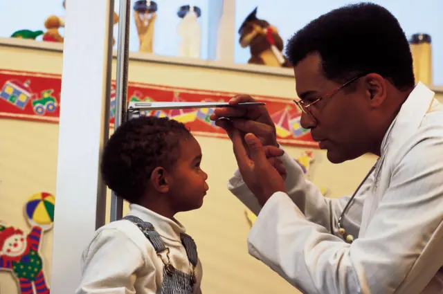 ਇੱਕ ਛੋਟਾ ਬੱਚਾ ਕੰਧ 'ਤੇ ਲੱਗੇ ਇੱਕ ਕੱਦ ਮਾਪਣ ਵਾਲੇ ਵਰਟੀਕਲ ਸਕੇਲ ਕੋਲ ਸਿੱਧਾ ਖੜ੍ਹਾ ਹੈ। ਚਿੱਟੇ ਕੋਟ ਵਿੱਚ ਇੱਕ ਬਾਲਗ, ਬੱਚੇ ਦੇ ਕੋਲ ਖੜ੍ਹਾ ਹੈ ਅਤੇ ਉਸ ਨੇ ਬੱਚੇ ਦੇ ਸਿਰ ਉੱਤੇ ਇੱਕ ਮਾਪਣ ਵਾਲੀ ਲੇਟਵੀਂ ਪੇਟੀ ਰੱਖੀ ਹੋਈ ਹੈ, ਜੋ ਕੱਦ ਚੈੱਕ ਕਰਨ ਲਈ ਸਕੇਲ ਦੇ ਨਾਲ ਮਿਲਾਈ ਗਈ ਹੈ।