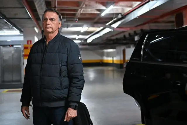 Bolsonaro sério, em pé, ao lado de carro em estacionamento