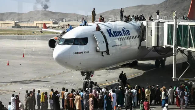 People climb atop a plane as they wait at the Kabul airport, and crowd around, there is smoke in the background