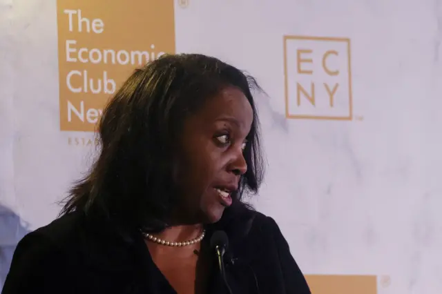 Lisa de perfil, falando algo em frente a painel de evento