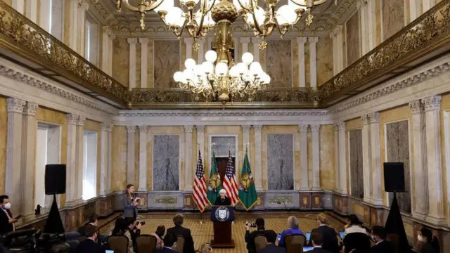 一間金碧輝煌的大廳內，時任美國財政部長珍尼特·耶倫（Janet Yellen）正在對觀眾發表演說。大廳四周矗立著新古典風格的柱子，牆面鑲嵌著大理石裝飾，中段則有一道鍍金的夾層陽台，懸掛在高聳的牆面之上。
