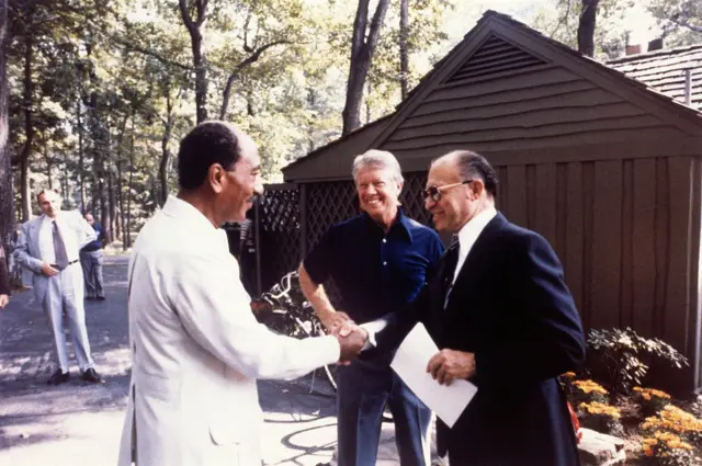 Anwar Sadat e Menachem Begin apertando as mãos, com Jimmy Carter ao fundo 