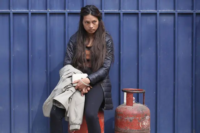 Seorang perempuan sedang duduk di atas gas LPG berwarna merah dengan wajah kesal sambil memangku jaketnya.