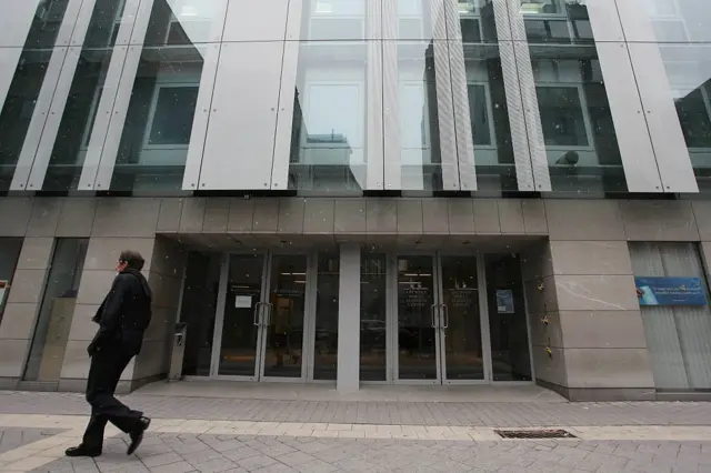 Fachada moderna de vidrio y concreto con varias puertas de entrada y una persona caminando por la acera