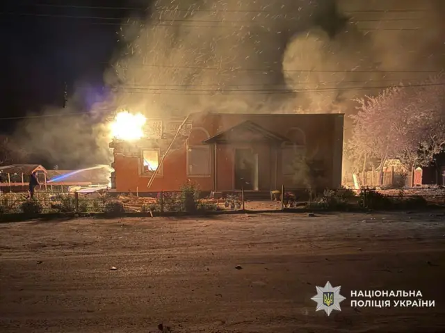 Горить помаранчевий будинок. Вогонь гасить пожежник, який стоїть від нього ліворуч