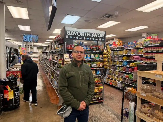Carlos Macias parado frente a los estantes de su pequeña tienda de comestibles. Tiene anteojos y viste una chaqueta verde oliva. Un cliente camina por una de las hileras del almacén