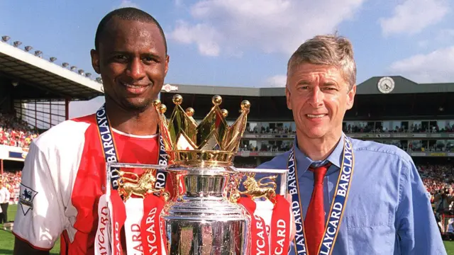 Patrick Vieira, vêtu d'un maillot rouge et blanc d'Arsenal, brandit le trophée de la Premier League orné de rubans rouges et blancs à hauteur de poitrine. Il se tient aux côtés d'Arsène Wenger, qui porte une chemise bleue et une cravate rouge. Tous deux, visibles jusqu'à la tête, ont des rubans bleus autour du cou, retenant des médailles hors champ. À l'arrière-plan, on aperçoit le Clock End du stade d'Highbury.
