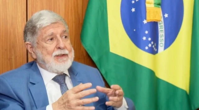 Foto de Celso Amorim gesticulando com as mãos e com uma bandeira do Brasil do lado direito da tela