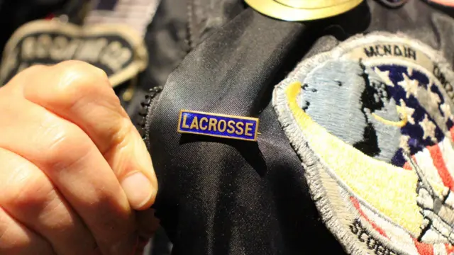 A close up of a small rectangular lacrosse badge being held towards the camera while pinned on Ace's jacket