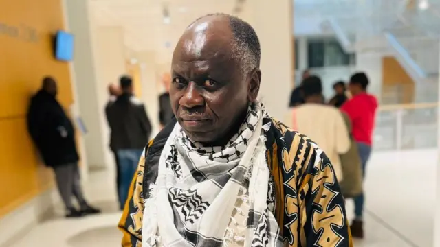 Nkiko Nsengimana ahagaze yiteye fulari mu ijosi, inyuma ye hari abandi bantu batagaragara neza 