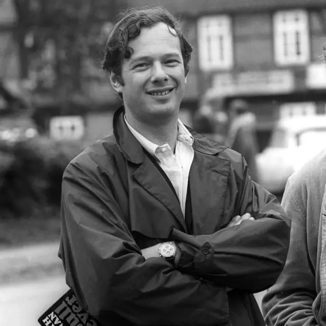 黑白照片：身穿斗篷大衣的爱波斯坦翘手展现笑容（9/1966）