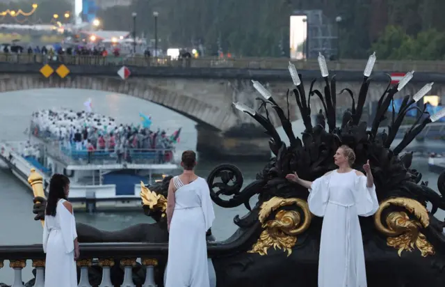 Des artistes vêtus de robes blanches se tiennent sur le Pont Alexandre III lors de la cérémonie d'ouverture des Jeux olympiques de Paris 2024, alors qu'un bateau passe en dessous.