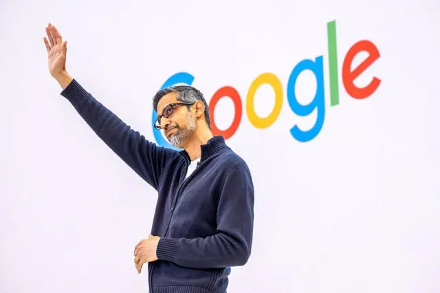 Сундар Пичаи, CEO компаний Alphabet и Google, стоит на сцене на фоне логотипа Гугл и поднимает в приветствии руку